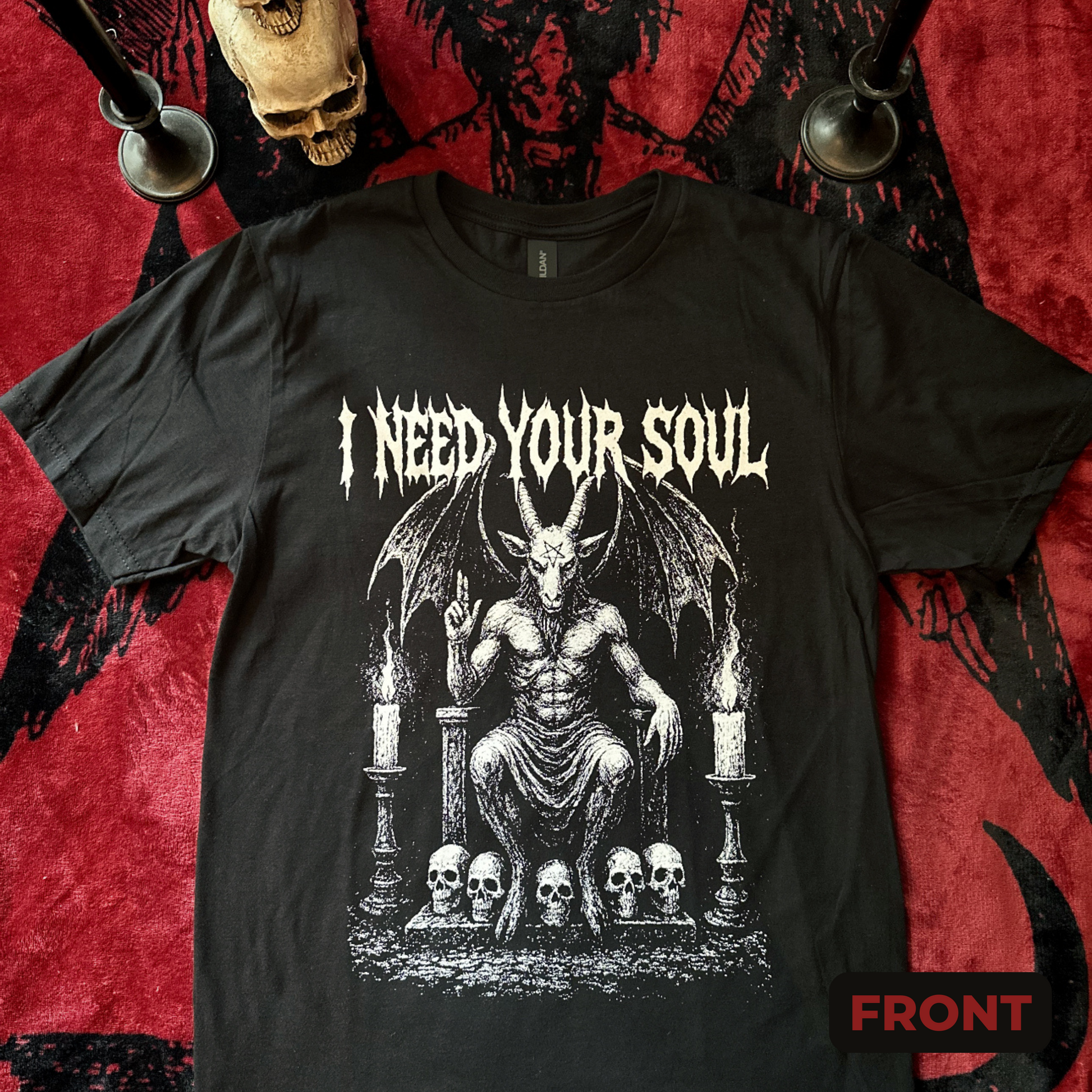 This black t-shirt showcases a striking graphic of a horned, winged figure seated on a throne, flanked by burning candles and resting upon a row of skulls